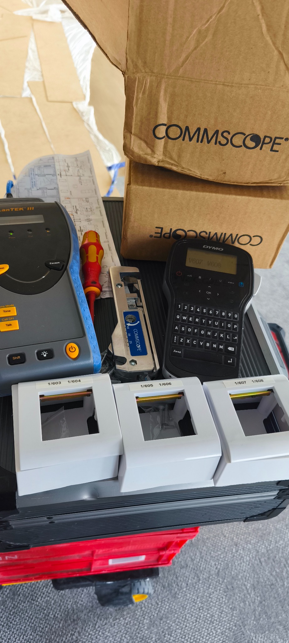 Tester kabli LanTEK III, etykietownica Dymo, zaciskarka CommScope i moduły keystone z oznaczeniami, ułożone na kufrze narzędziowym. Widoczny plan instalacji.