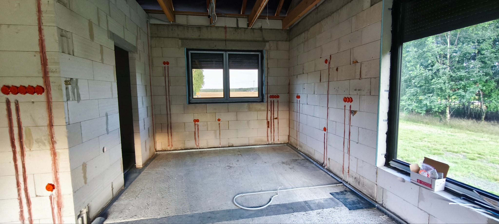 Wnętrze w trakcie instalacji elektrycznej: ściany z betonu komórkowego z puszkami i przewodami, okno z roletą zewnętrzną, widoczne elementy instalacji podłogowej.