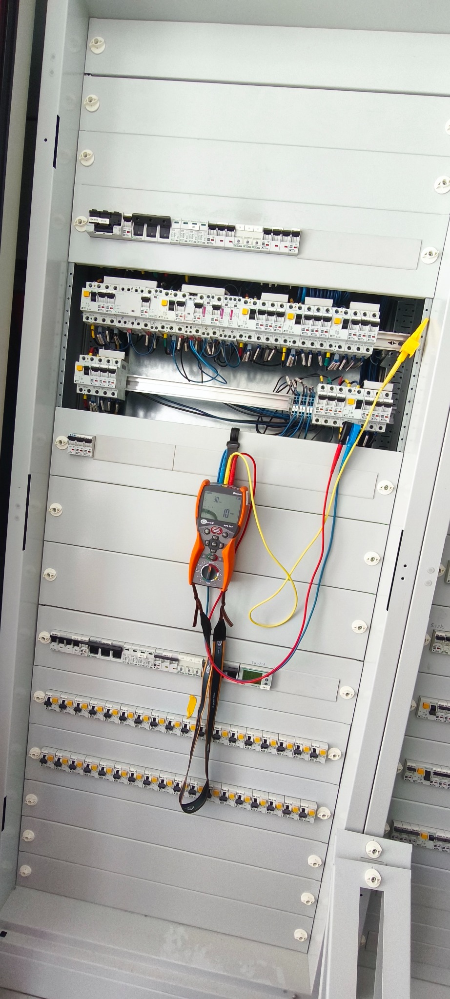 Otwarta szafa elektryczna z zamontowanymi bezpiecznikami i przewodami. Podłączony multimetr cyfrowy wskazuje pomiar. Widoczne kable w kolorach żółtym, czerwonym i niebieskim.