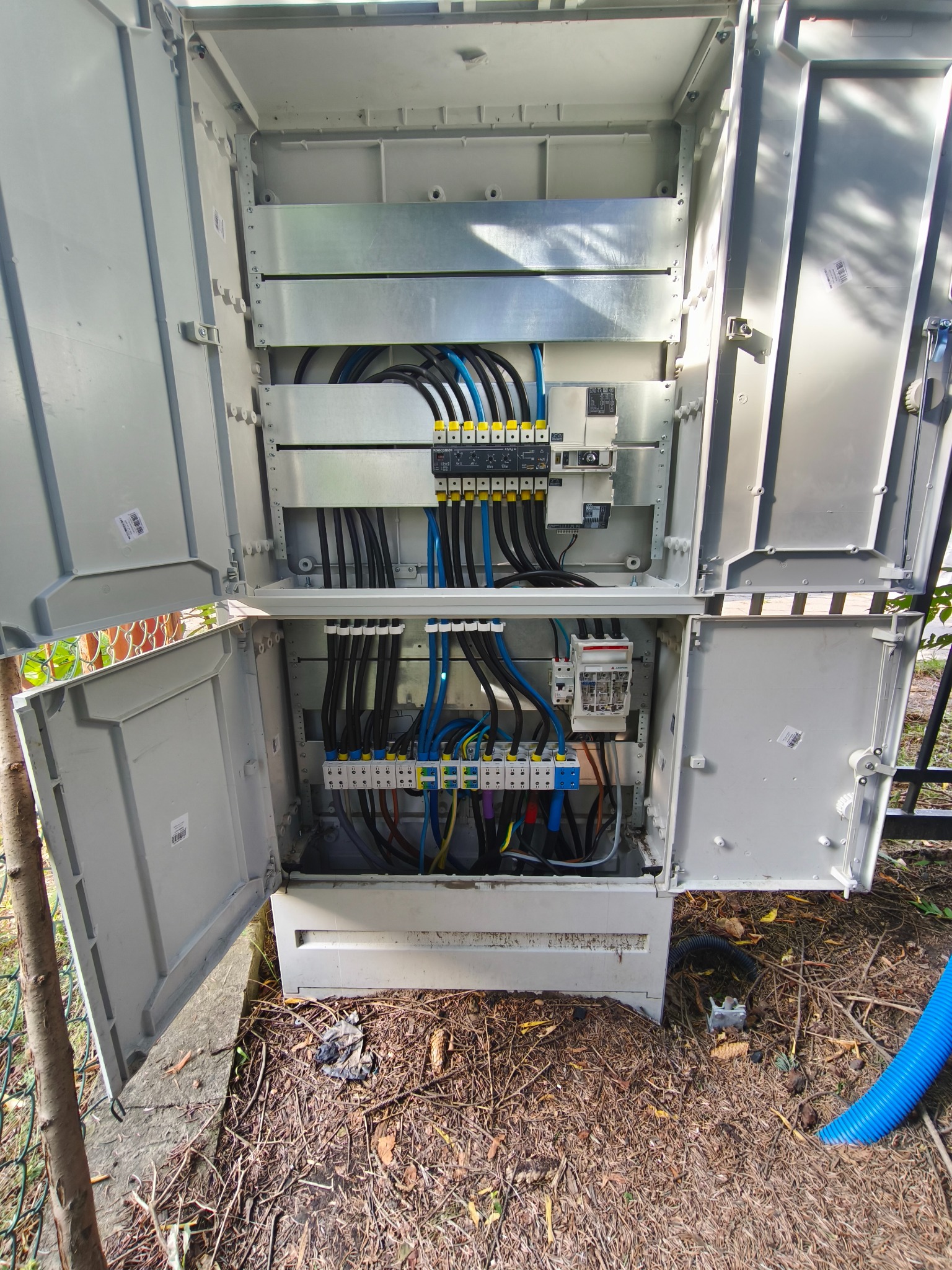 Otwarta, szara skrzynka elektryczna z widocznymi kablami i bezpiecznikami, umieszczona na zewnątrz, na tle ziemi z liśćmi i fragmentem niebieskiego węża.