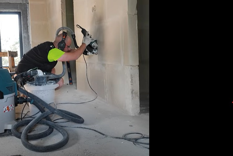 Pracownik w słuchawkach tnie ścianę z betonu komórkowego szlifierką kątową z odkurzaczem przemysłowym. Widoczne przewody i poziomica.