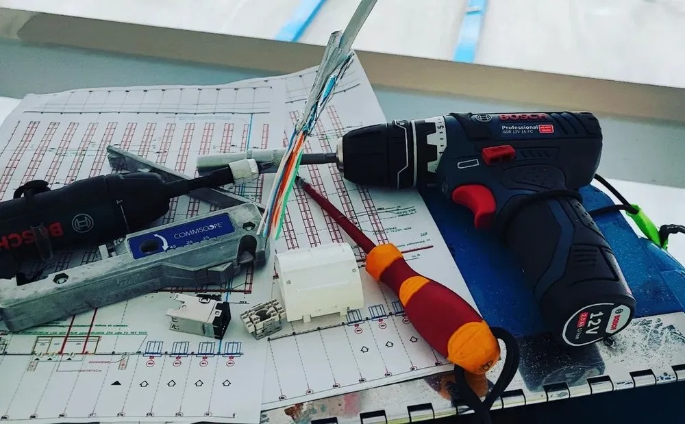 Narzędzia i schemat instalacji: wiertarka Bosch, zaciskarka Commscope, wkrętak, kabel sieciowy i gniazdo na planie instalacji. Prace instalacyjne w toku.