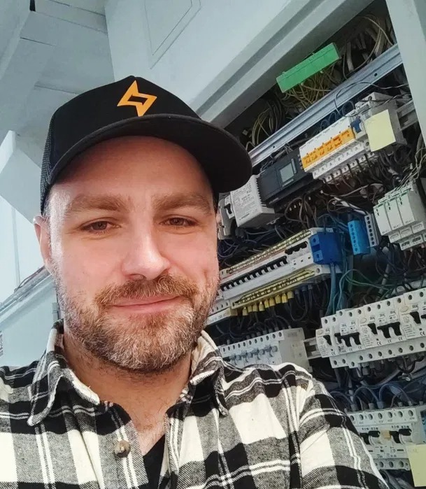 Uśmiechnięty elektryk w czapce z logo na tle rozdzielnicy z bezpiecznikami i okablowaniem. Ujęcie z bliska, widoczna twarz i górna część korpusu.