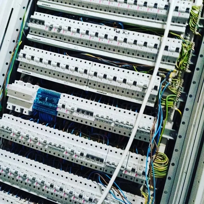 Rozdzielnia elektryczna z zamontowanymi bezpiecznikami i okablowaniem, widok z bliska, ukośne ujęcie. Elementy instalacji w kolorze białym, niebieskim, zielonym i żółtym.