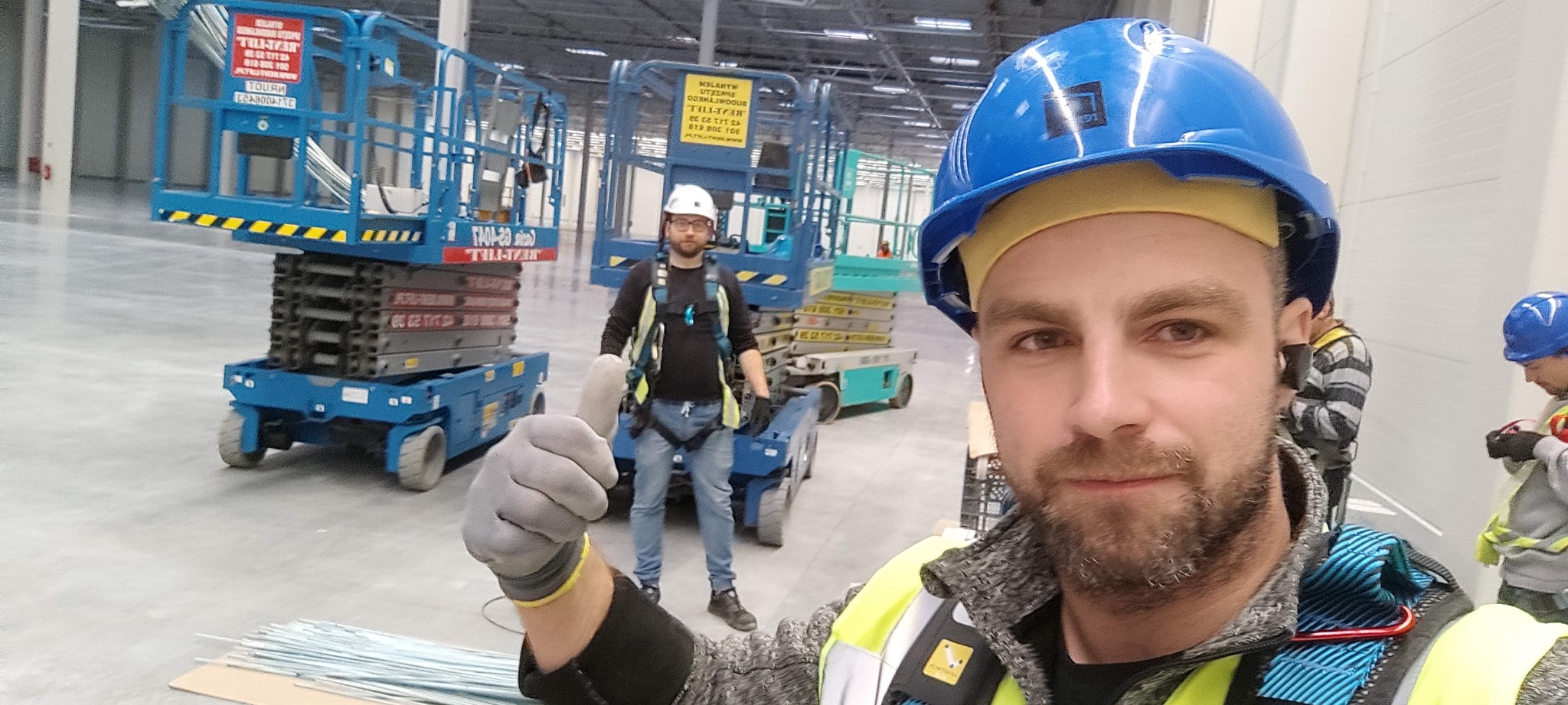 Instalator w kasku, z uśmiechem, na tle hali z podnośnikami i współpracownikami. Praca zespołowa przy instalacjach elektrycznych w obiekcie przemysłowym.