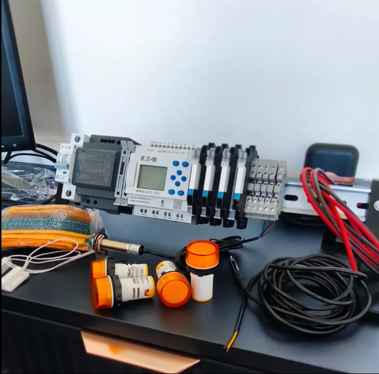 Panel sterowania Eaton z modułami, czujnikami i przewodami na czarnym blacie. Widoczny fragment monitora. Elementy instalacji elektrycznej.