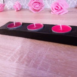 Drewniany, czarny świecznik na trzy tealighty z różowymi wkładami, ustawiony na blacie z różowego drewna, w tle dekoracja z różowych róż.