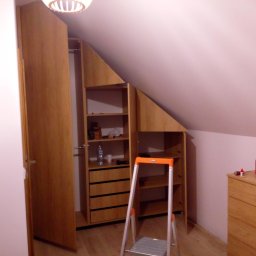 Drewniana szafa wnękowa na poddaszu, z otwartymi drzwiami i drabinką. Widoczne półki i szuflady. Ustawiona pod skosem dachu. Na komodzie obok pudełko chusteczek.