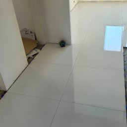 REMONT HARANT - Białe, błyszczące płytki podłogowe ułożone w jasnym pomieszczeniu. Widoczny laser krzyżowy i niebieskie klipsy poziomujące. Częściowo widoczne okno.