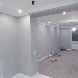 REMONT HARANT - Wykończone wnętrze w odcieniach szarości z dekoracyjną sztukaterią sufitową i nowoczesnym oświetleniem punktowym. Na podłodze widoczne narzędzia pracy.