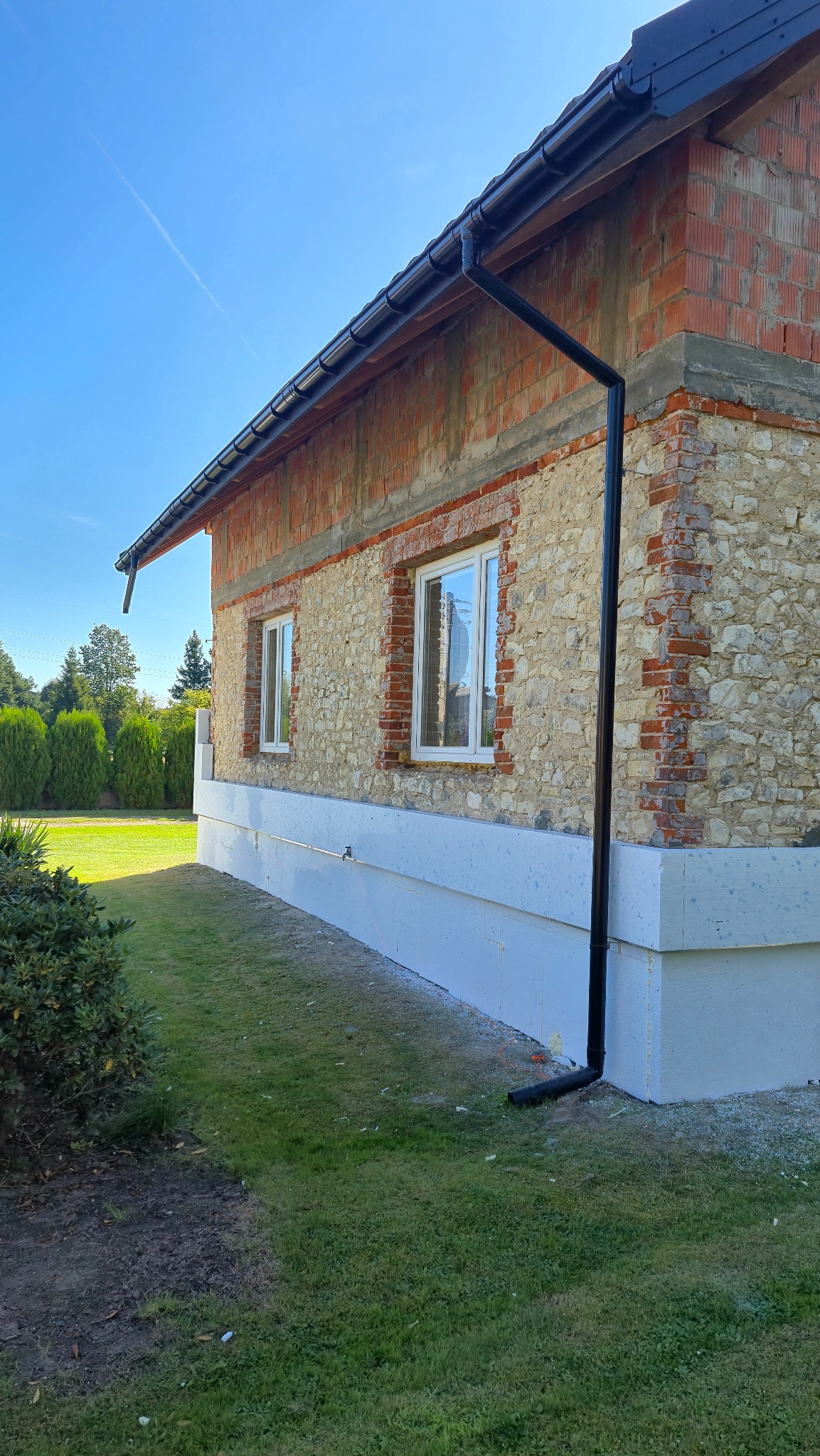 Dom w trakcie renowacji: kamienna elewacja z ceglanymi obramowaniami okien, ocieplony fundament i czarna rynna. Widok z zewnątrz na tle zieleni.