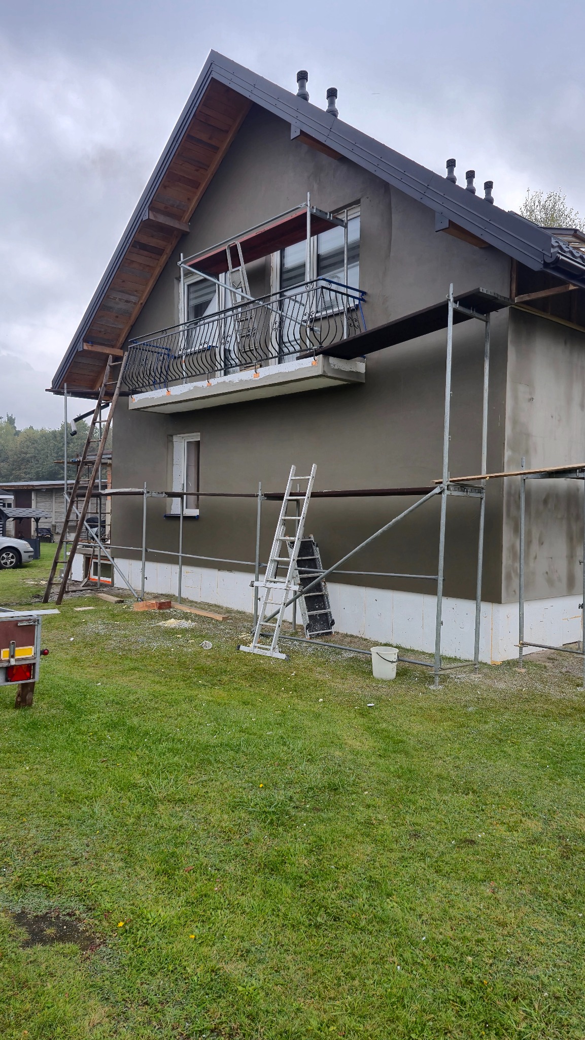 Dom jednorodzinny w trakcie prac elewacyjnych, widoczne rusztowania, drabiny i świeżo nałożony tynk w szarym odcieniu. Balustrada balkonowa z metaloplastyki.