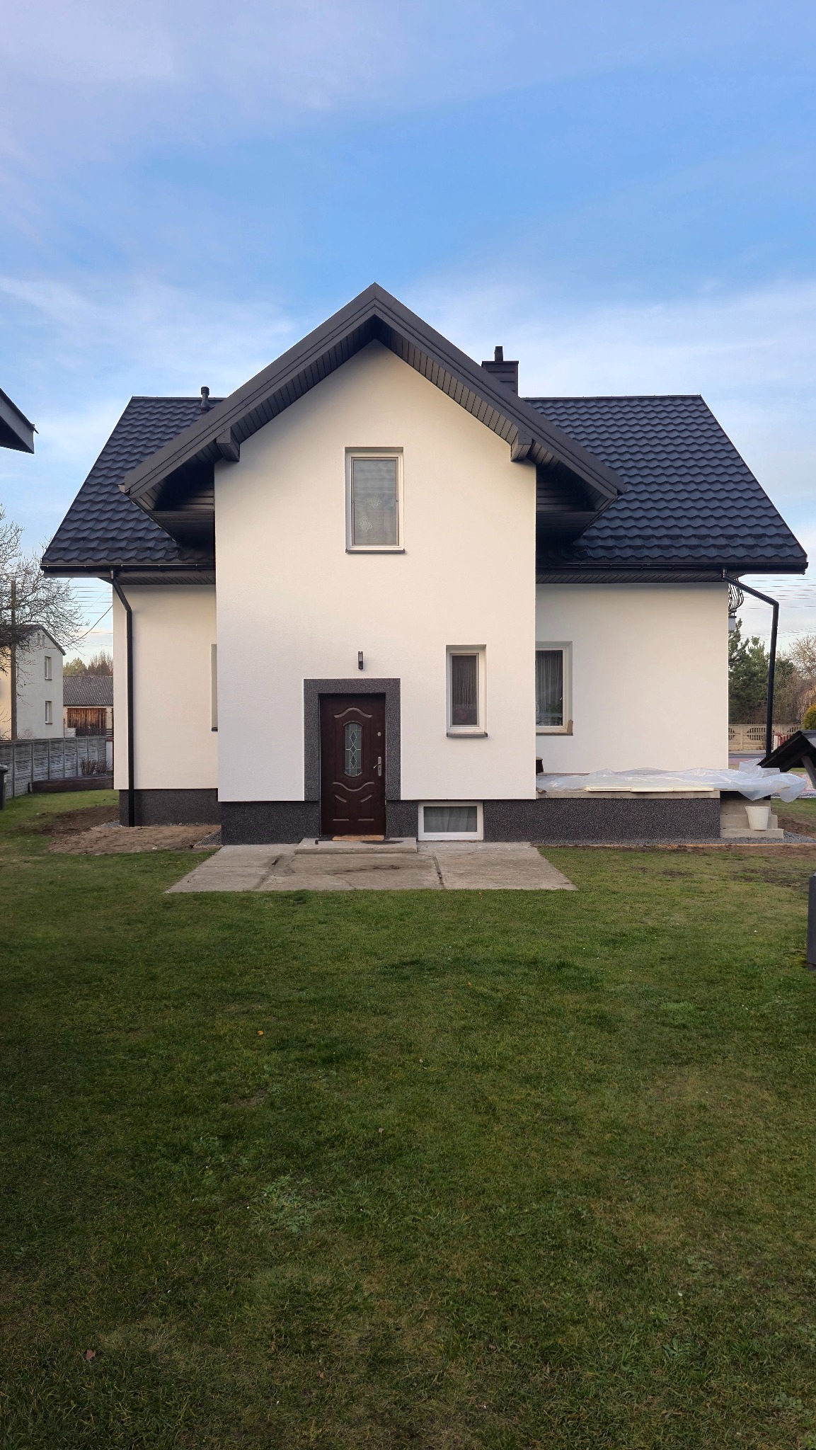 Biały dom jednorodzinny z ciemnym dachem i stolarką okienną, widok z trawnika. Elewacja po remoncie, z widocznymi detalami wykończenia.
