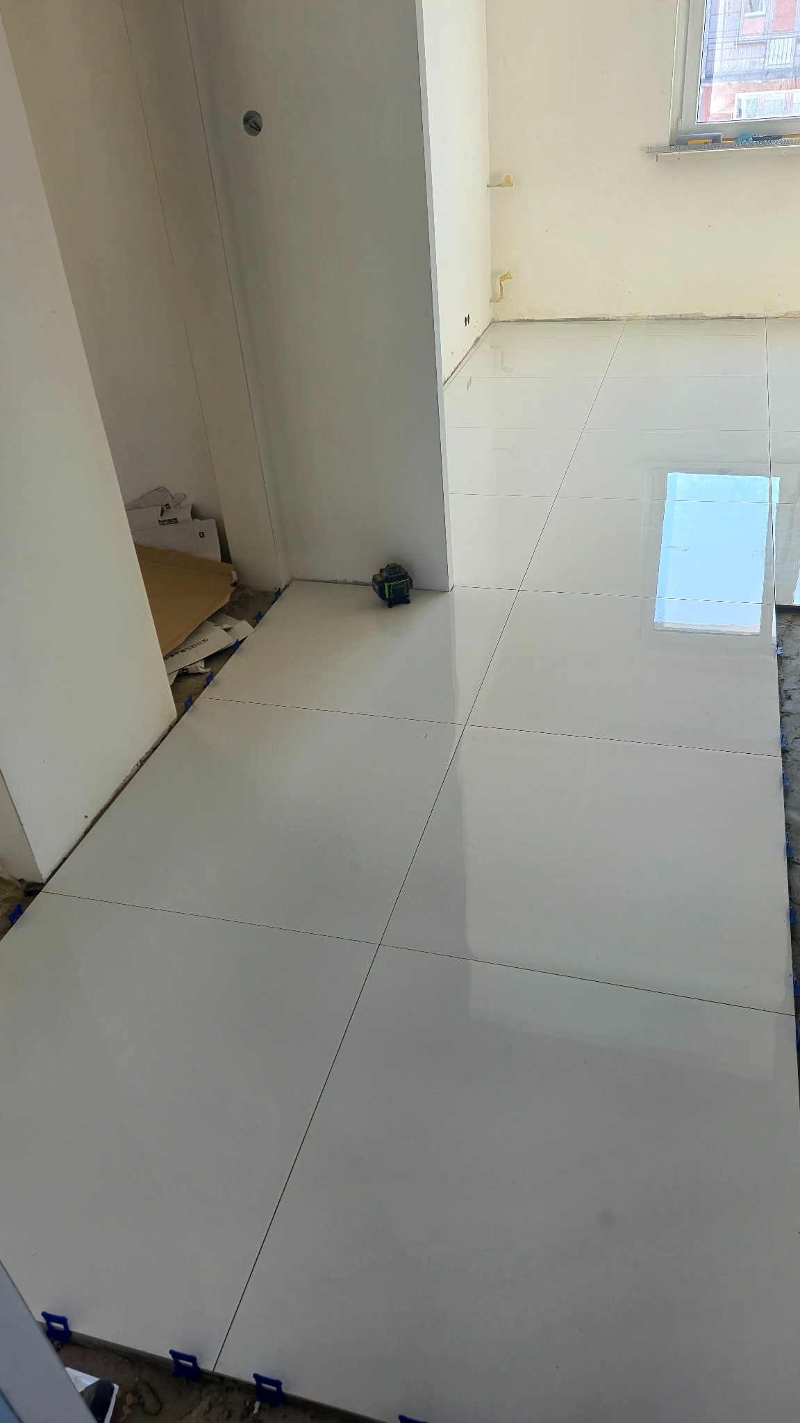 Białe, błyszczące płytki podłogowe ułożone w jasnym pomieszczeniu. Widoczny laser krzyżowy i niebieskie klipsy poziomujące. Częściowo widoczne okno.