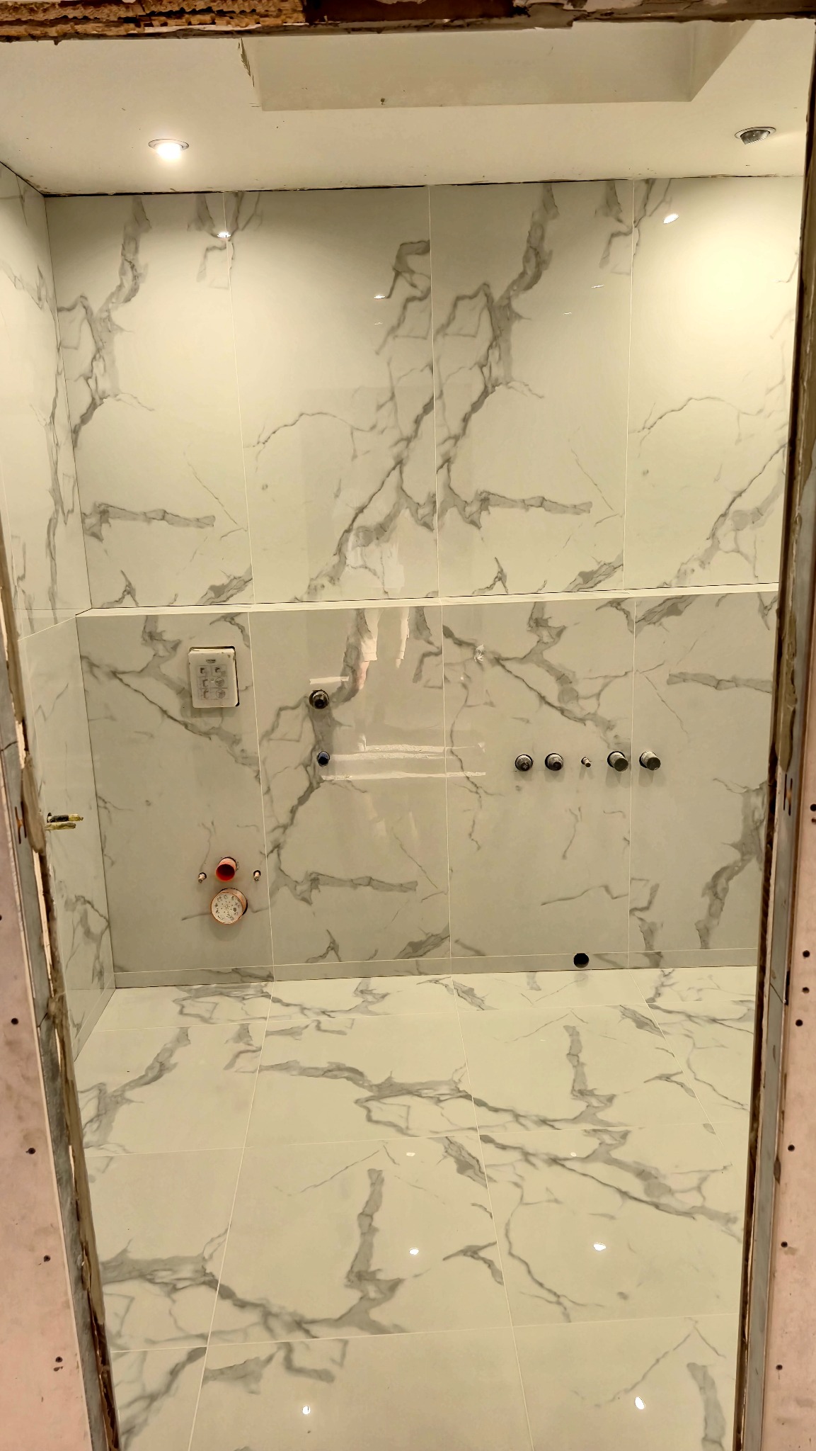 Wykończona łazienka z białymi płytkami imitującymi marmur, widok z perspektywy wejścia, armatura zamontowana na ścianie, oświetlenie punktowe.