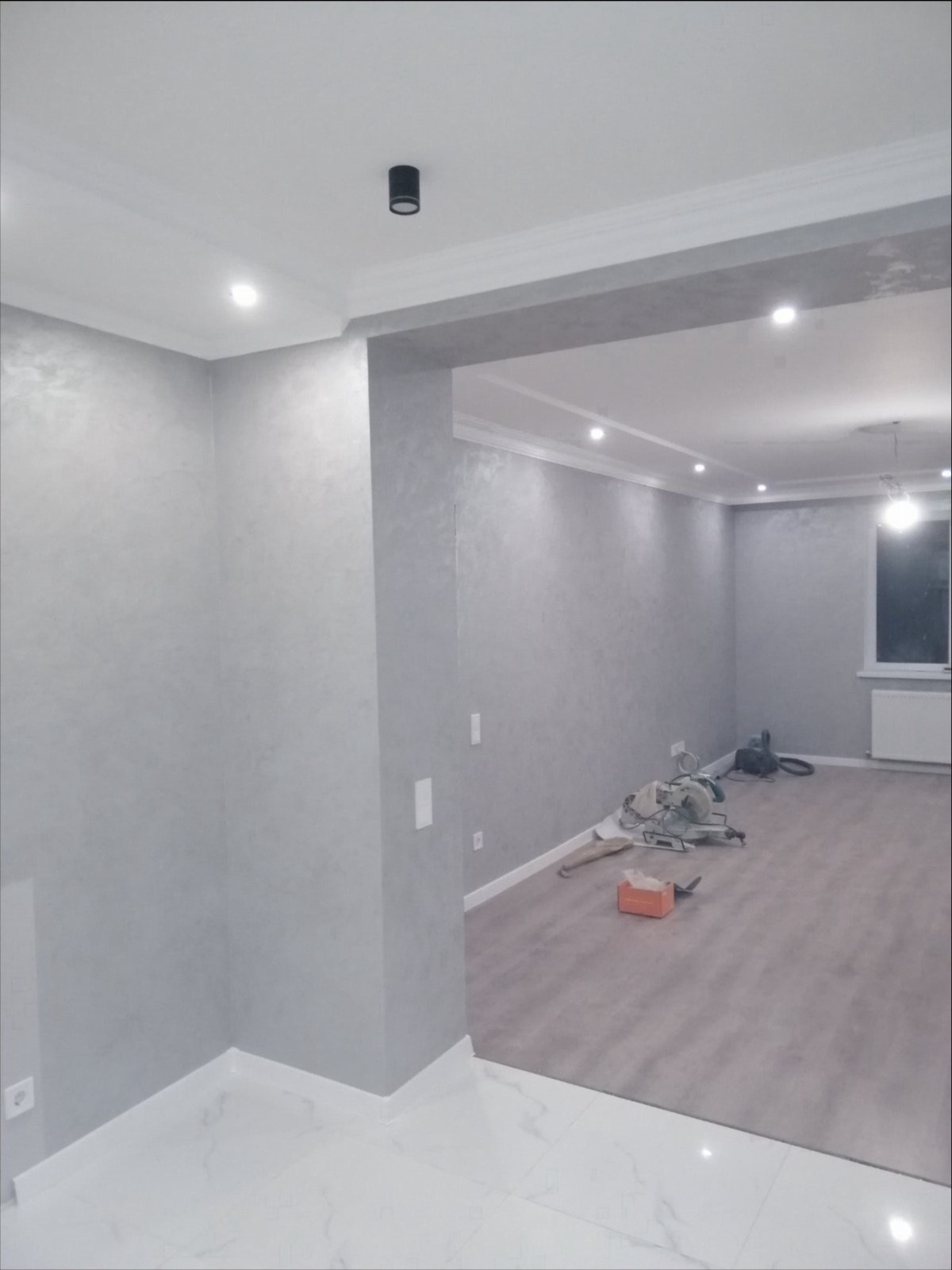 Wykończone wnętrze w odcieniach szarości z dekoracyjną sztukaterią sufitową i nowoczesnym oświetleniem punktowym. Na podłodze widoczne narzędzia pracy.