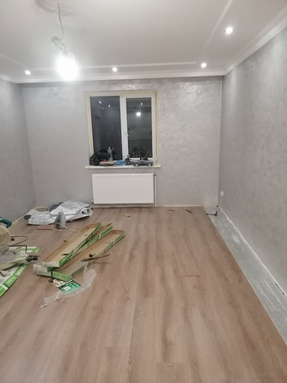 Układanie paneli podłogowych w pokoju z oknem i grzejnikiem. Widoczne narzędzia i materiały budowlane. Ściany w odcieniach szarości z dekoracyjną fakturą.