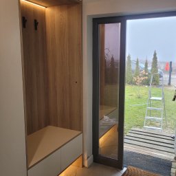 Anstol - Nowoczesna szafa wnękowa z oświetleniem LED w przedpokoju, widok na zewnątrz przez szklane drzwi. Minimalistyczny design, jasne kolory, drewnopodobne wykończenie.