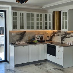 Anstol - Biała kuchnia z przeszklonymi szafkami górnymi i złotymi akcentami, marmurowy backsplash, złoty okap, widok na zewnątrz przez okno. Nowoczesny design.