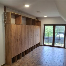 Anstol - Długa szafa z drewnianymi frontami i otwartymi półkami u dołu, zamontowana w pokoju na poddaszu z oknem balkonowym i podłogą imitującą drewno.