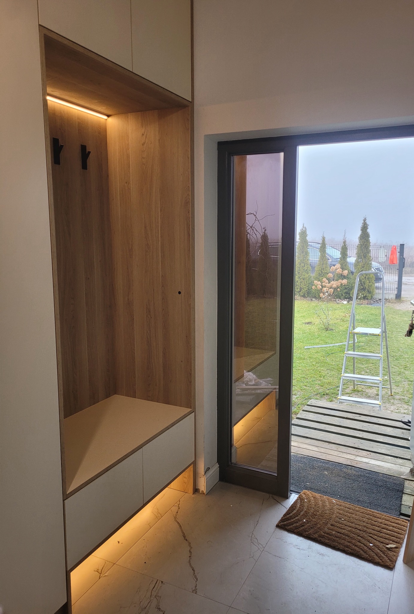 Nowoczesna szafa wnękowa z oświetleniem LED w przedpokoju, widok na zewnątrz przez szklane drzwi. Minimalistyczny design, jasne kolory, drewnopodobne wykończenie.