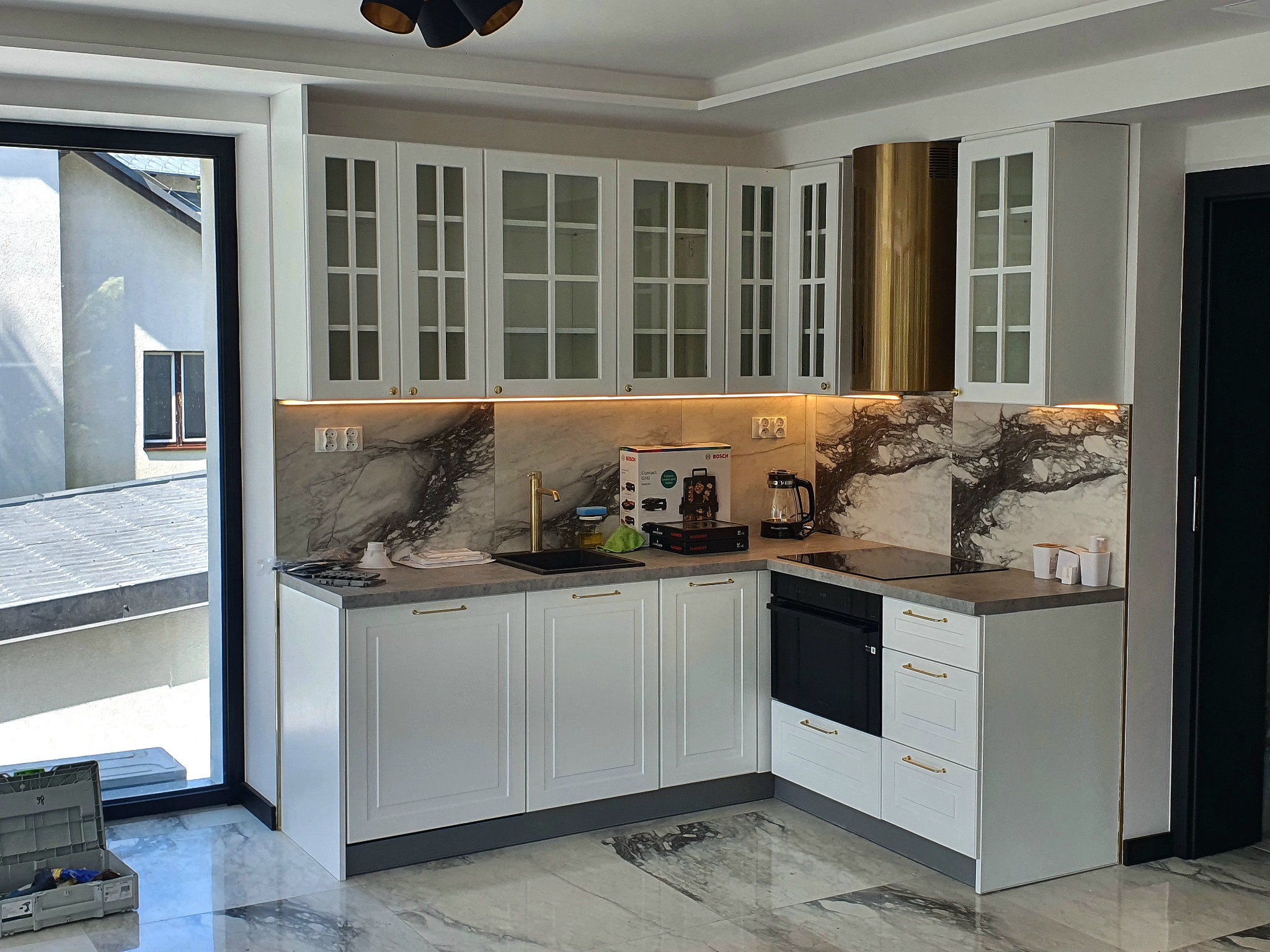 Biała kuchnia z przeszklonymi szafkami górnymi i złotymi akcentami, marmurowy backsplash, złoty okap, widok na zewnątrz przez okno. Nowoczesny design.