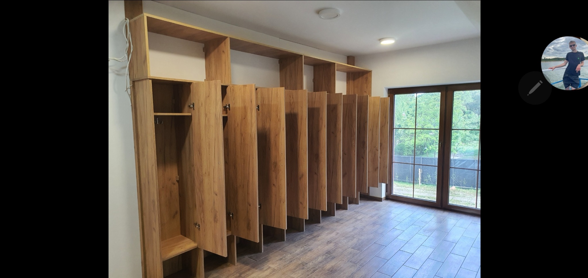 Długa zabudowa z szaf wnękowych z drewnianymi frontami, z otwartymi i zamkniętymi przestrzeniami, w jasnym pomieszczeniu z oknem na ogród.