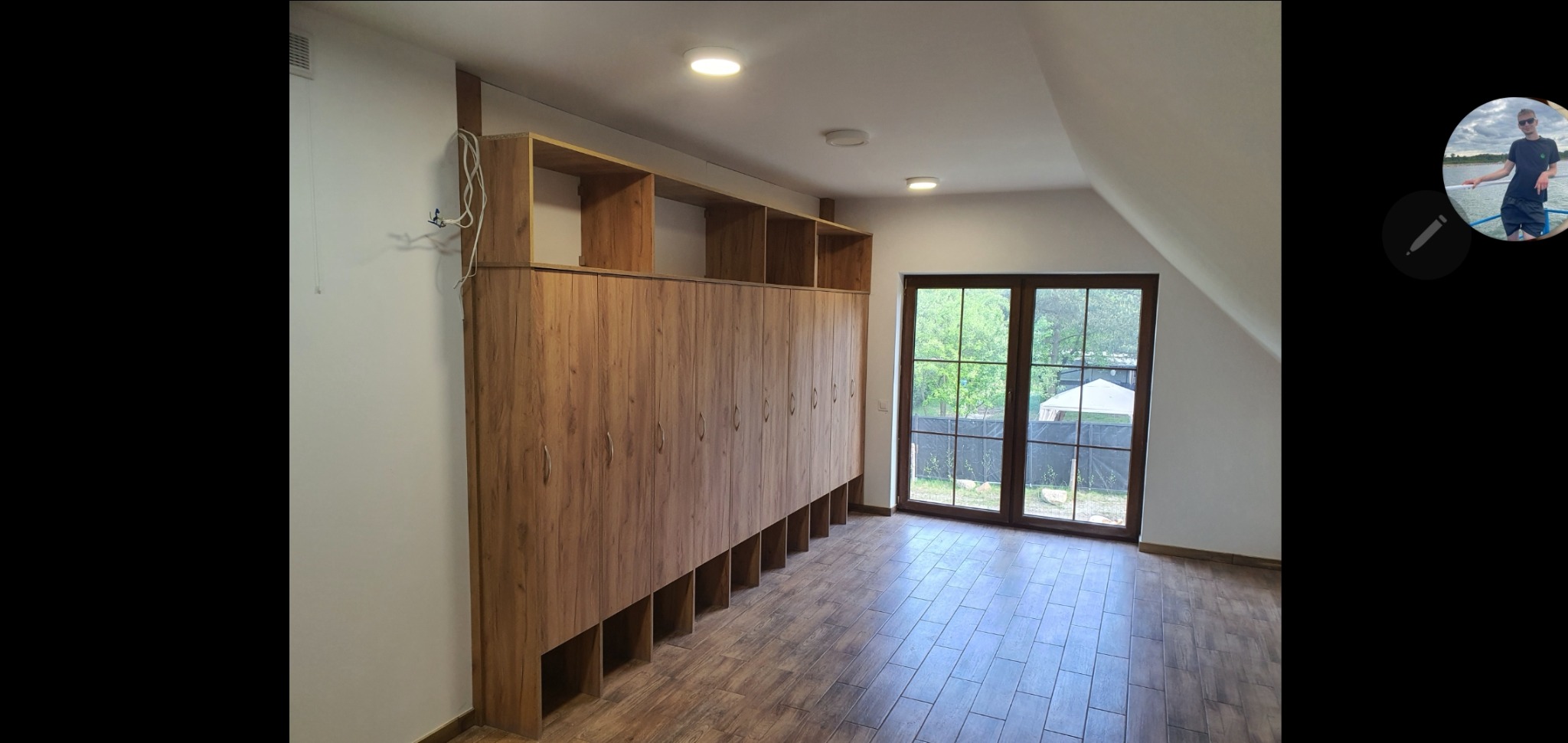 Długa szafa z drewnianymi frontami i otwartymi półkami u dołu, zamontowana w pokoju na poddaszu z oknem balkonowym i podłogą imitującą drewno.