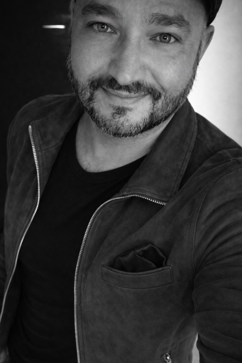 Czarno-biały portret uśmiechniętego mężczyzny z brodą w kurtce zamszowej, wykonany w Bydgoszczy. Ujęcie z góry, z widoczną górną częścią ciała.