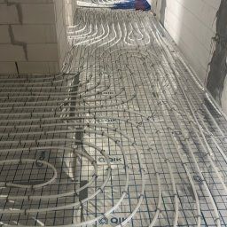 Therm-system - Białe rury ogrzewania podłogowego ułożone na folii z siatką w surowym wnętrzu budynku. Widoczne fragmenty ścian z białego pustaka i przygotowane podłoże.