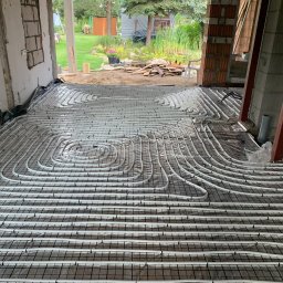 Therm-system - Białe rury ogrzewania podłogowego ułożone na czarnej siatce w surowym wnętrzu budynku w trakcie budowy. Widok z góry na instalację przed wylaniem posadzki.