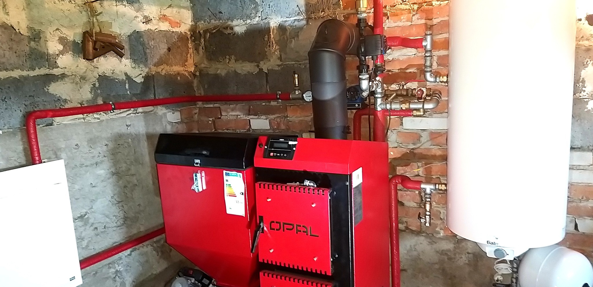 Czerwony piec CO marki Opal z czarnym kominem i białym zasobnikiem wody, zainstalowany w kotłowni z ceglanymi i betonowymi ścianami oraz czerwonymi rurami.