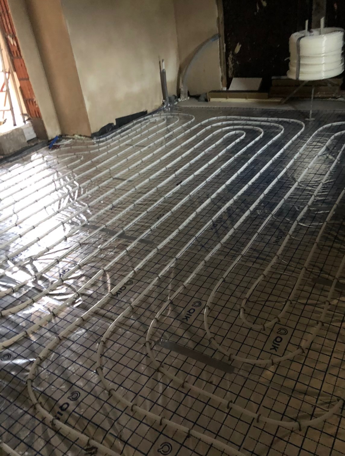 Białe rury ogrzewania podłogowego ułożone na folii z siatką w surowym wnętrzu. Widoczny fragment ściany z cegły i rolka rur. Etap instalacji przed wylewką.