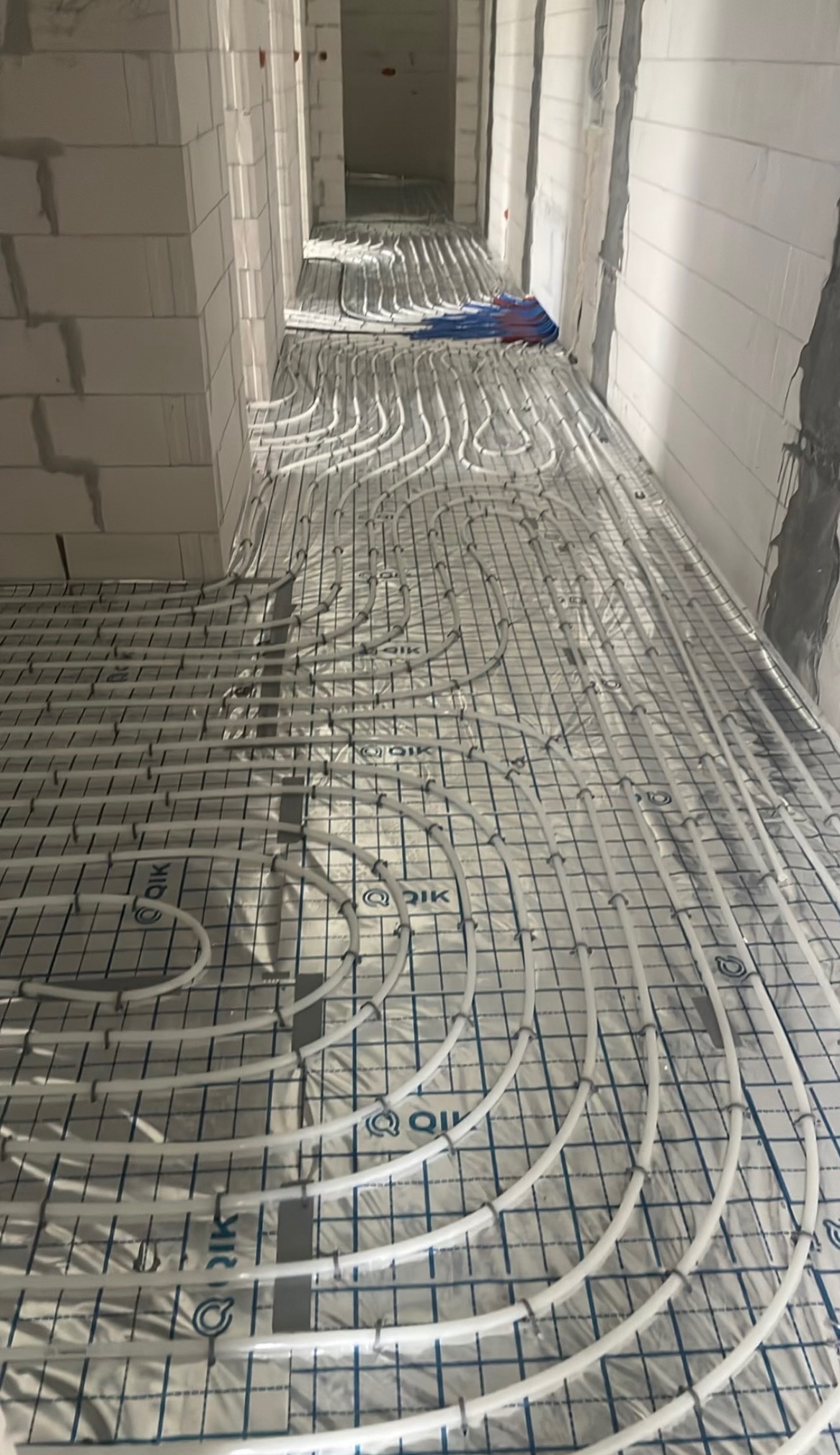 Białe rury ogrzewania podłogowego ułożone na folii z siatką w surowym wnętrzu budynku. Widoczne fragmenty ścian z białego pustaka i przygotowane podłoże.