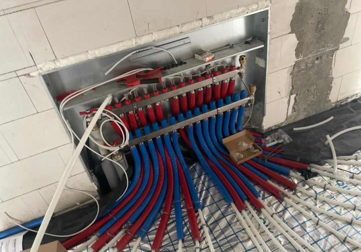 Rozdzielacz ogrzewania podłogowego z rurami w kolorze czerwonym i niebieskim, zamontowany w metalowej obudowie w surowym stanie wykończenia wnętrza.