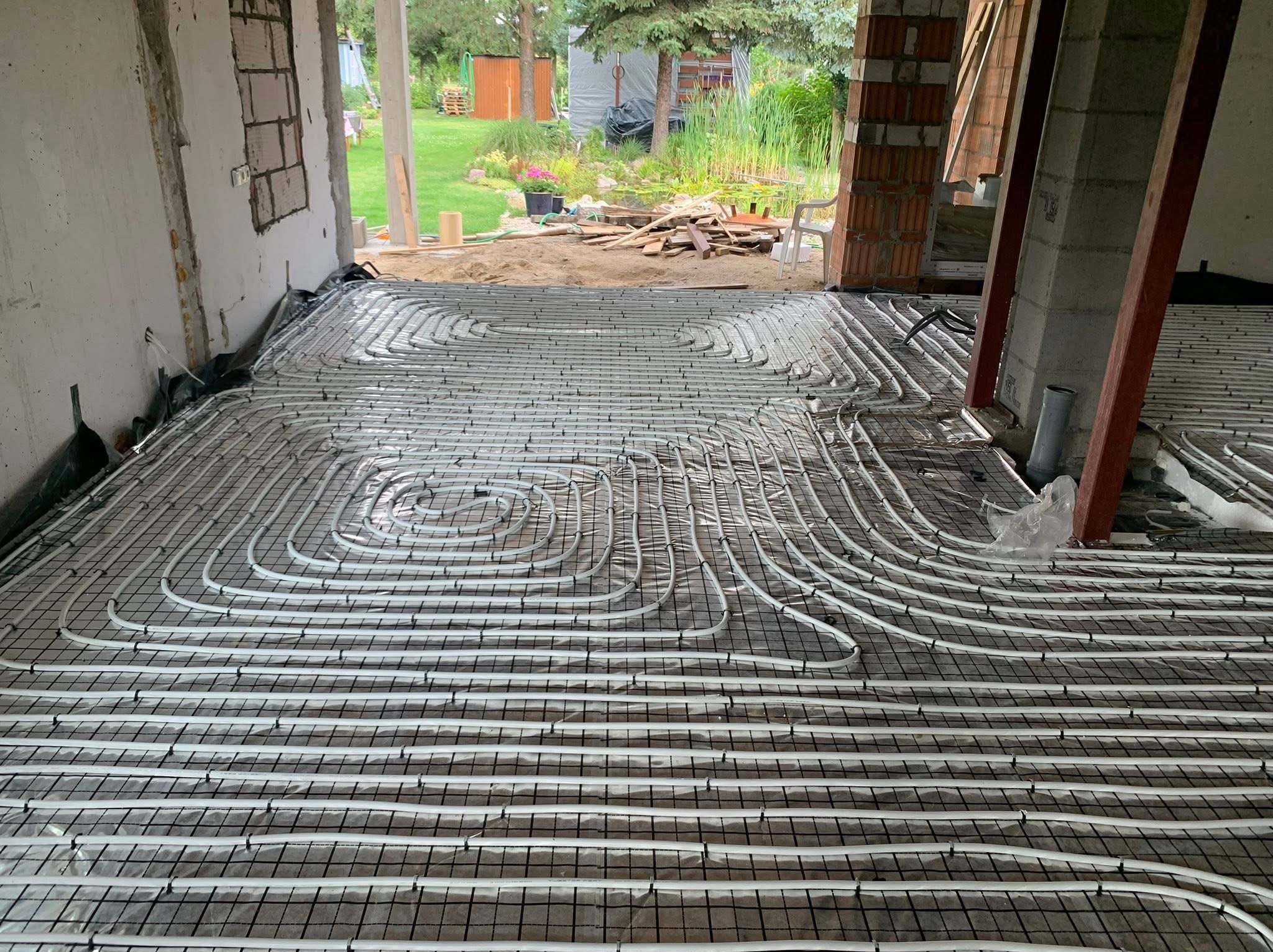 Białe rury ogrzewania podłogowego ułożone na czarnej siatce w surowym wnętrzu budynku w trakcie budowy. Widok z góry na instalację przed wylaniem posadzki.