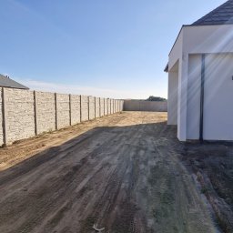 Grunt i Zieleń Piotr Jakubowski - Długa, prosta, szara, betonowa ścieżka z wyraźnymi śladami opon prowadzi wzdłuż jasnego, panelowego ogrodzenia do nowoczesnego, białego domu z szarym dachem.