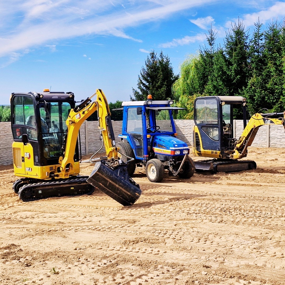 Dwa żółte mini-koparki CAT i niebieski traktor na piaszczystym terenie, ogrodzenie z betonowych elementów w tle, słoneczny dzień.
