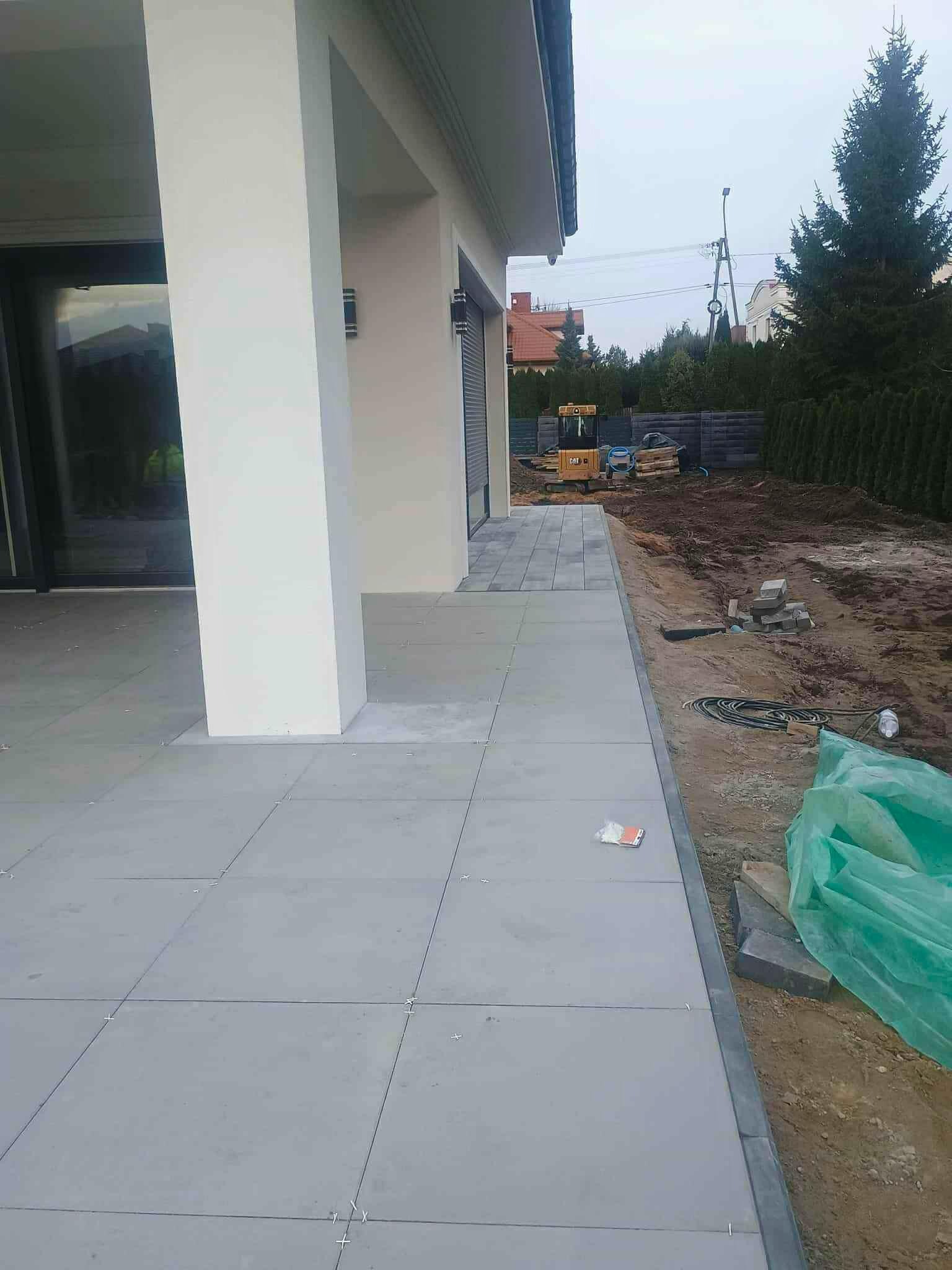 Układanie szarej kostki brukowej na posesji domu jednorodzinnego. W tle widoczny mini-ładowarka CAT i przygotowany teren pod dalsze prace. Widok z boku.
