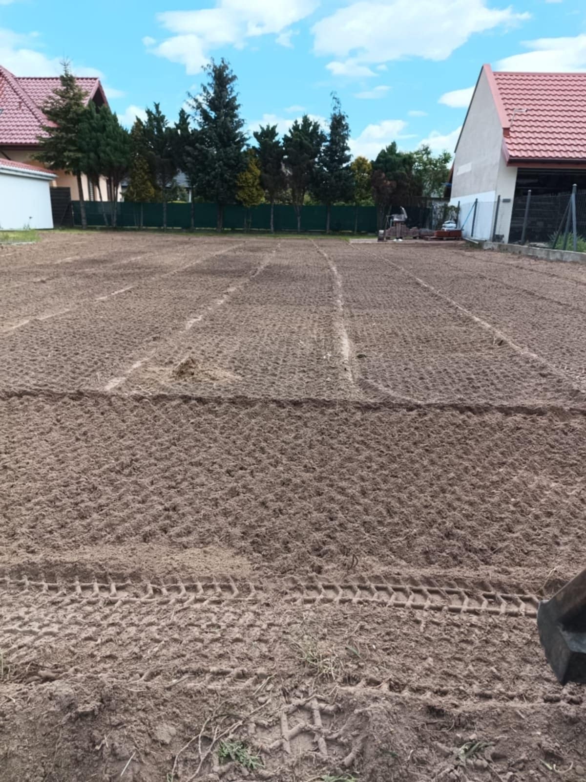 Przygotowany, wyrównany teren pod trawnik z widocznymi śladami opon, domy w tle i błękitne niebo. W oddali widoczna osoba pracująca na terenie.