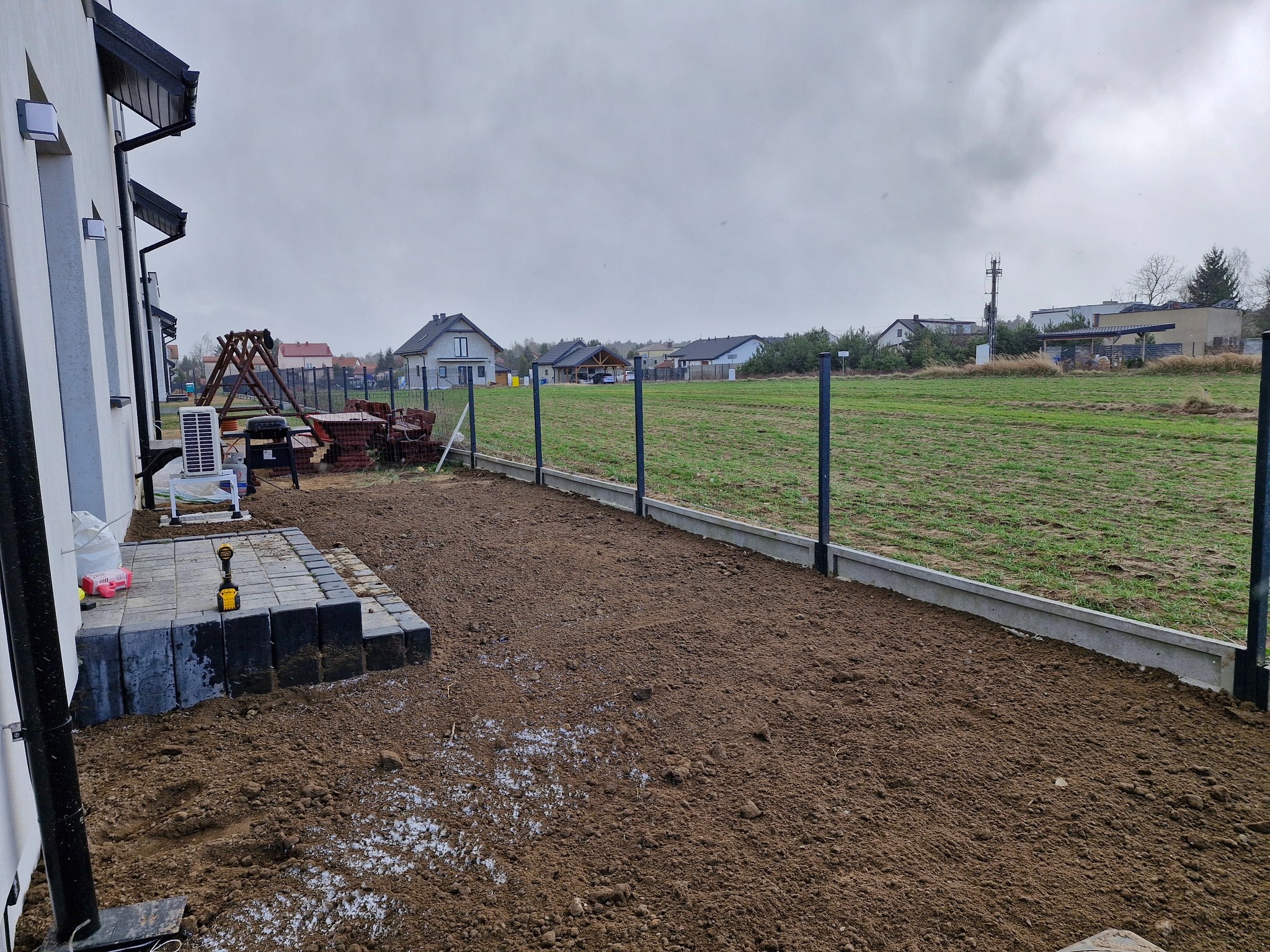 Przygotowany teren pod trawnik z częściowo ułożoną kostką brukową przy domu jednorodzinnym. Widoczne narzędzia i ogrodzenie panelowe. Pochmurne niebo.