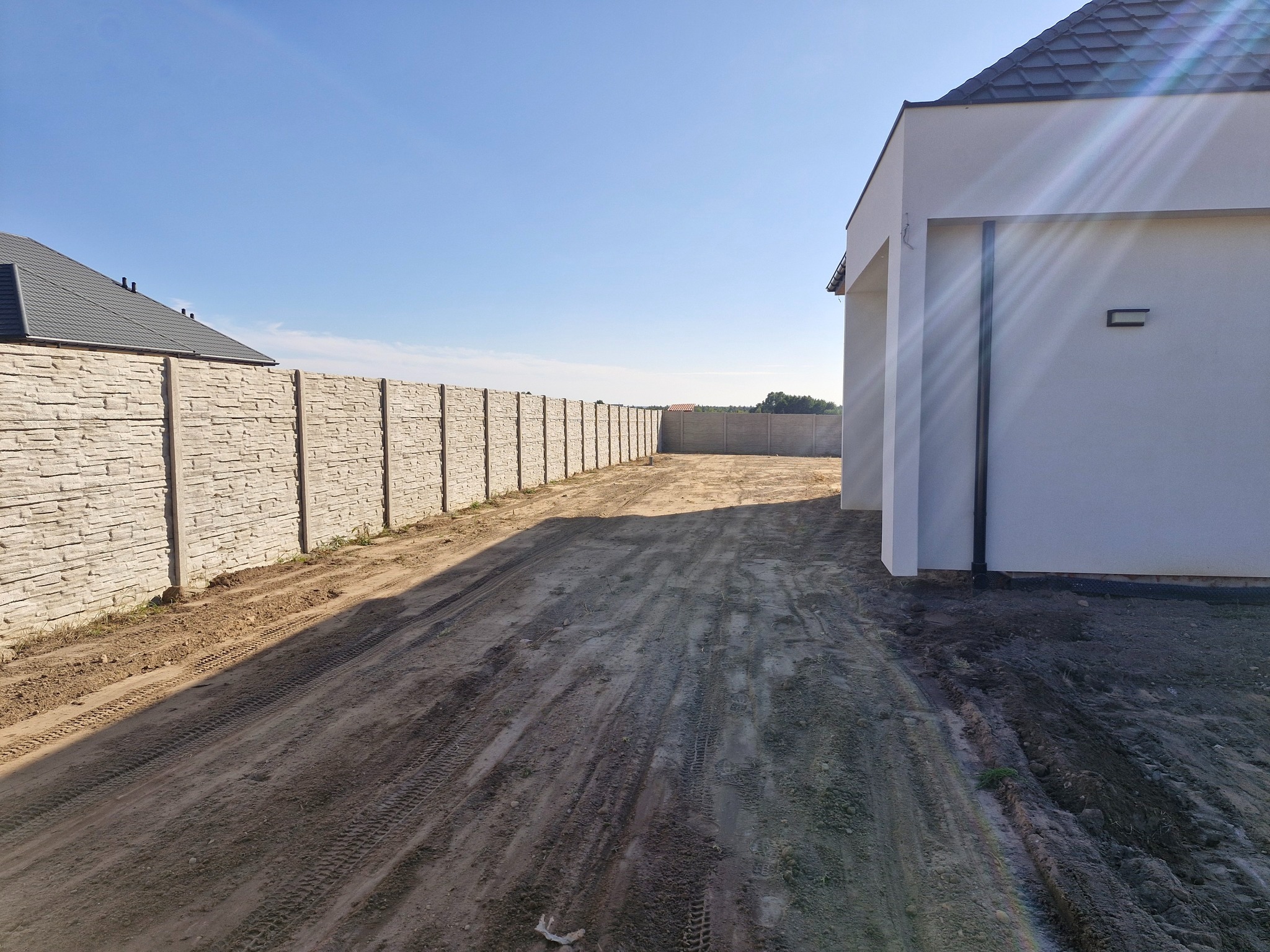 Długa, prosta, szara, betonowa ścieżka z wyraźnymi śladami opon prowadzi wzdłuż jasnego, panelowego ogrodzenia do nowoczesnego, białego domu z szarym dachem.