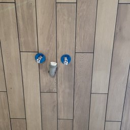 SOLID KAFEL POLAK - Ściana wyłożona podłużnymi, drewnopodobnymi płytkami z widocznymi przyłączami wodnymi i odpływem, ukazująca detale wykończenia w łazience.