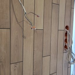 SOLID KAFEL POLAK - Ściana wyłożona podłużnymi płytkami imitującymi drewno, z widocznymi przewodami elektrycznymi i puszkami instalacyjnymi. Układ pionowy, wzór cegiełki.