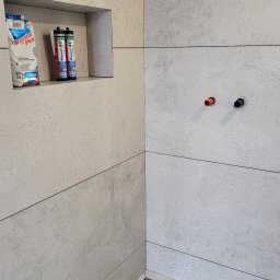 SOLID KAFEL POLAK - Wykończona ściana w łazience z szarych, matowych płytek imitujących kamień. Wnęka z chemią budowlaną i odpływ liniowy w podłodze. Widoczne przyłącza wodne.