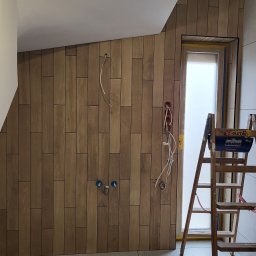 SOLID KAFEL POLAK - Ściana wyłożona drewnopodobnymi płytkami w łazience, widoczne wyprowadzenia hydrauliczne i elektryczne, drabina Krause. Ukończony etap prac.