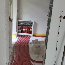 Solid Bud - Czerwone rury ogrzewania podłogowego ułożone na posadzce, obok biały rozdzielacz z czerwonymi zaworami i pojemnik Ceresit, w remontowanym wnętrzu.