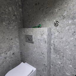 Solid Bud - Nowoczesna łazienka z podwieszaną miską WC, wykończona szarymi płytkami lastryko-podobnymi. Na zabudowie leży zielone narzędzie, obok wystają przewody elektryczne.