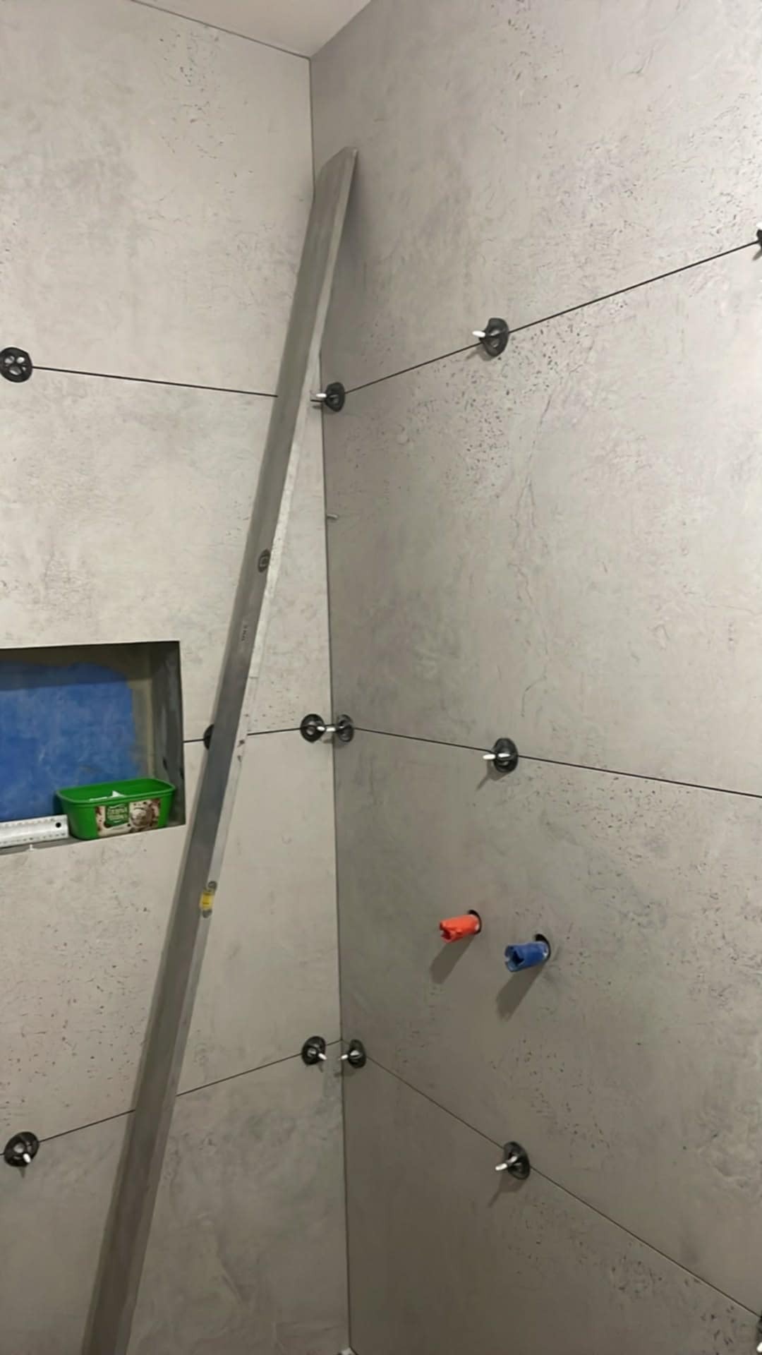 Ściana łazienki w trakcie wykańczania, z szarymi płytkami, poziomica oparta o ścianę, widoczne elementy instalacji wodnej i system poziomowania płytek.
