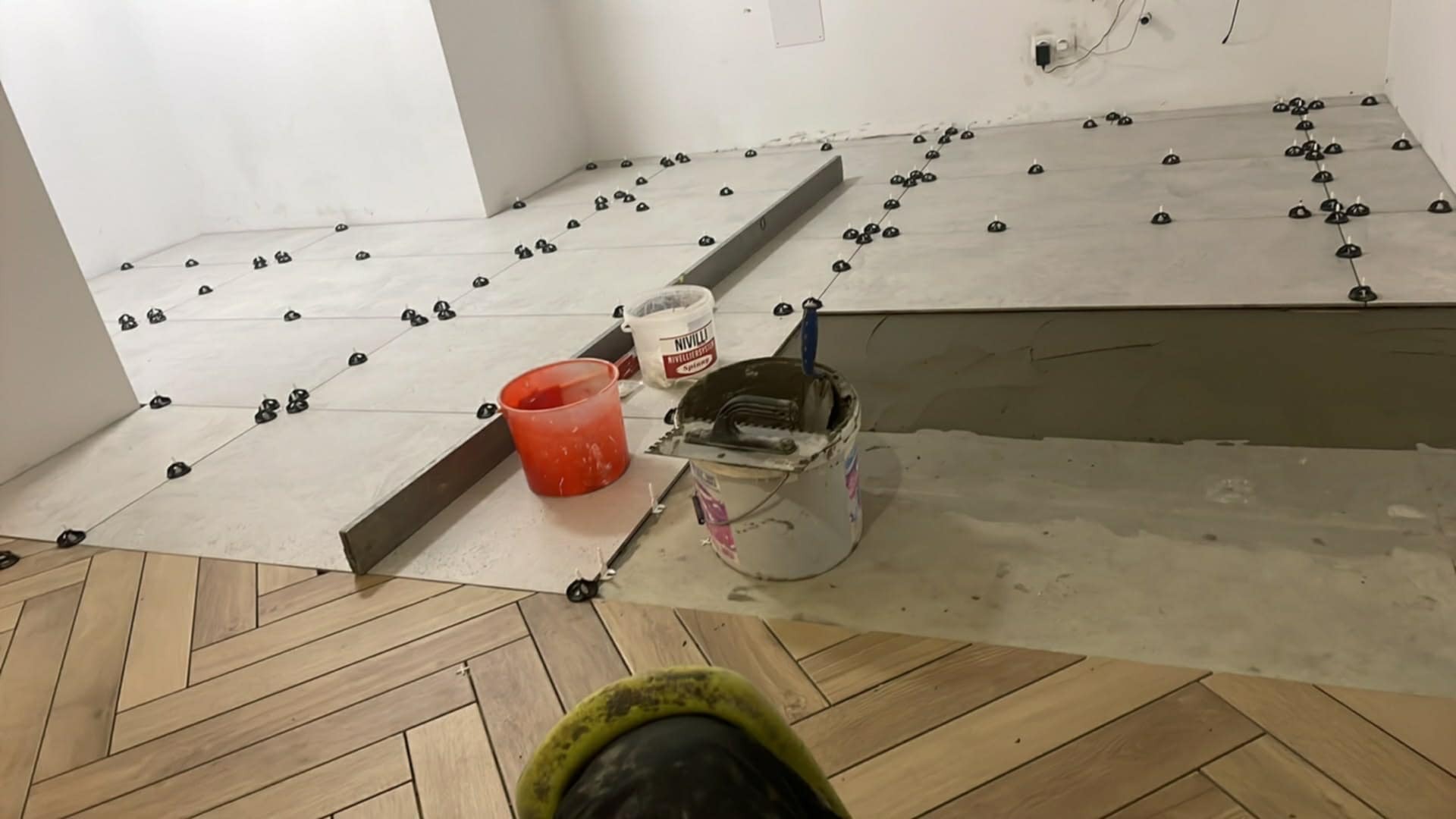 Etap prac: układanie jasnych płytek z użyciem systemu poziomowania na podłodze, z fragmentem parkietu w jodełkę. Widoczne narzędzia i materiały budowlane.